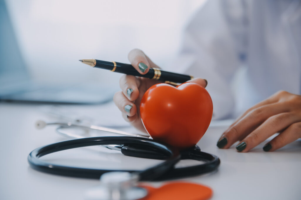 Medico con camice e stetoscopio tiene una penna accanto a un cuore di plastica sul tavolo