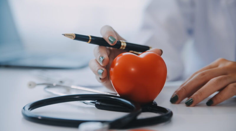 Medico con camice e stetoscopio tiene una penna accanto a un cuore di plastica sul tavolo