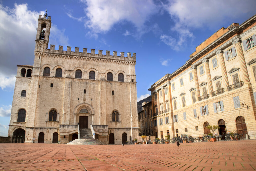 Piazza medievale di Gubbio con edifici storici in pietra, caffè all’aperto e atmosfera tipica umbra”